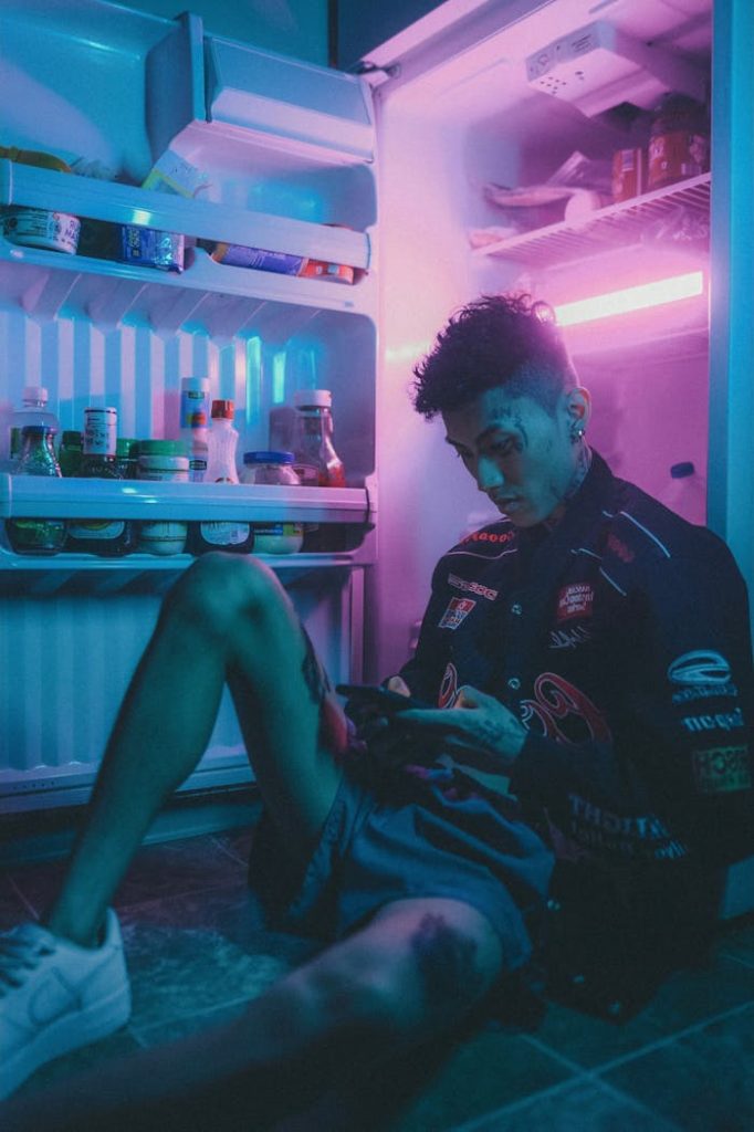 Tattooed man uses phone while sitting beside neon-lit open fridge.